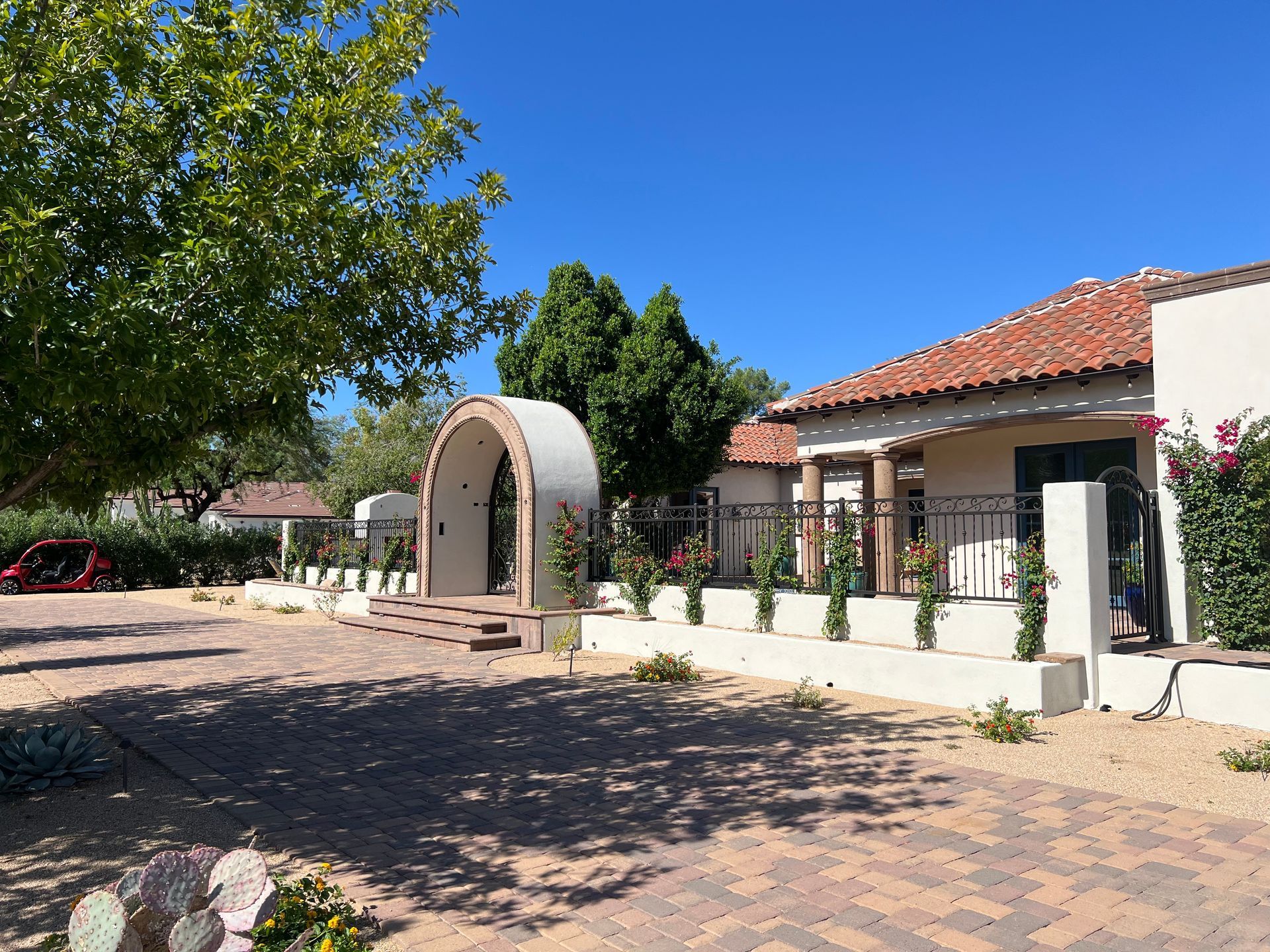 Sunny courtyard with a tiled-roof building, arched entryway, and trees casting shadows on a cobblestone path