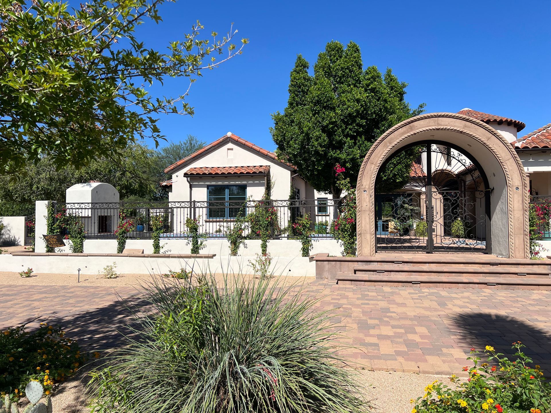 Sunny courtyard with a white stucco building, red tile roof, and arched brick walkway amid trees and shrubs