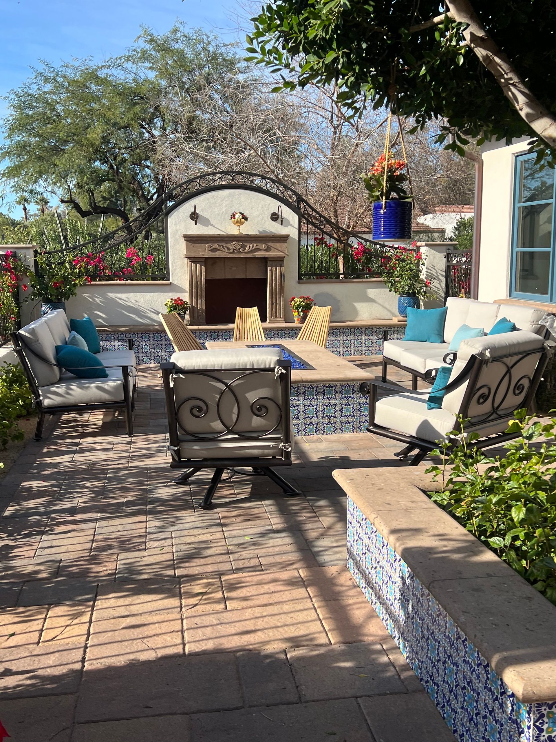 Outdoor patio with cushioned chairs, a table, and a small fireplace surrounded by colorful plants and brick pavers