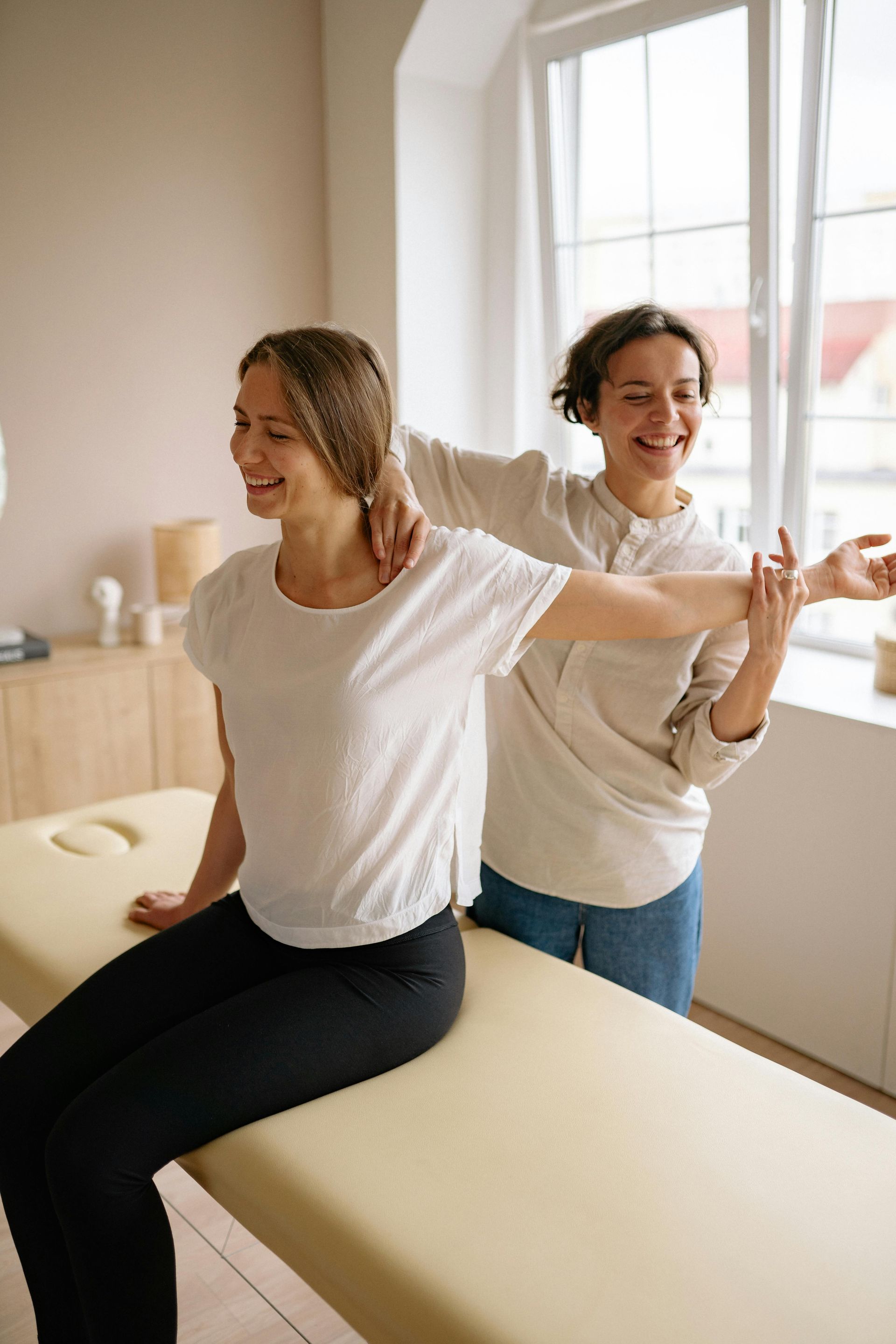 Two people smiling and stretching in a bright bedroom, one seated on the bed and one standing behind