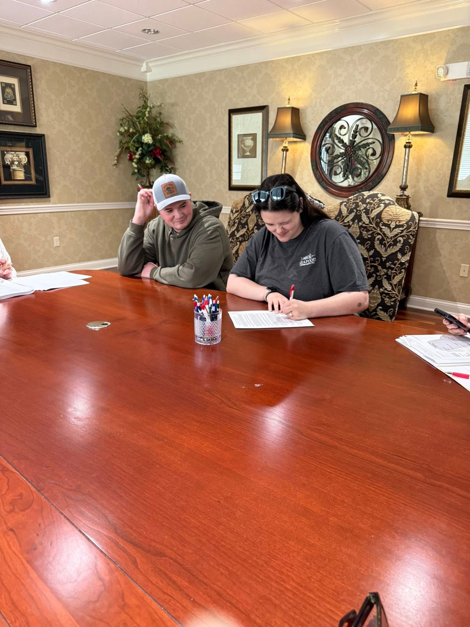 Two people seated at a large wooden conference table, writing on paper in a warmly lit room.