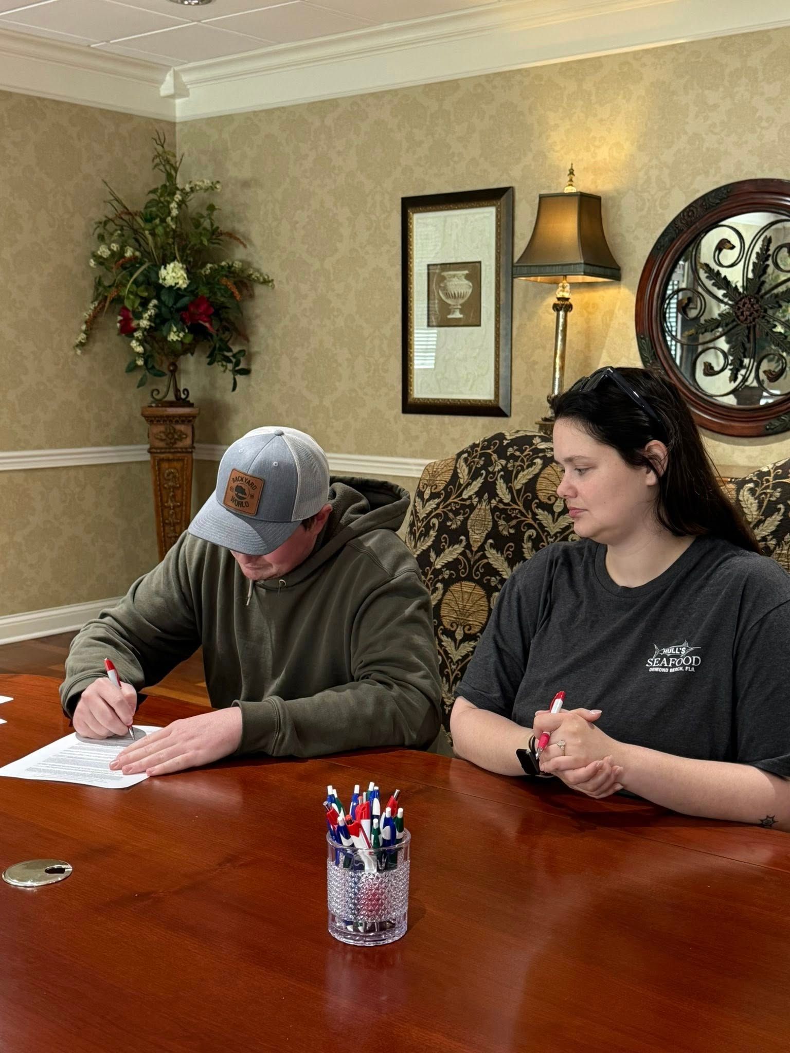 Two people seated at a dining table, one writing on paper while the other watches, in a living room.