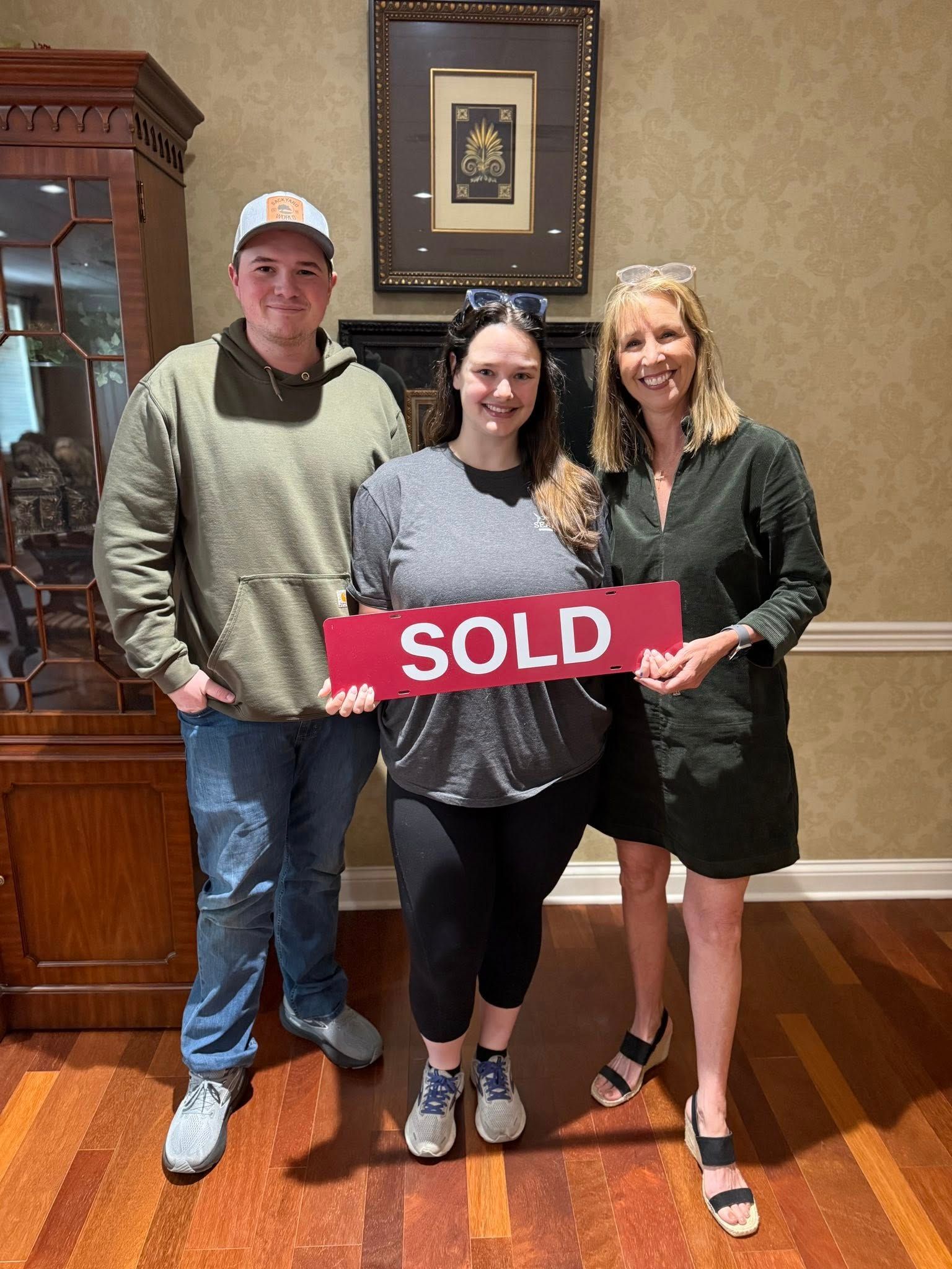 Three people indoors hold a red “SOLD” sign, standing on a hardwood floor with framed art behind them.