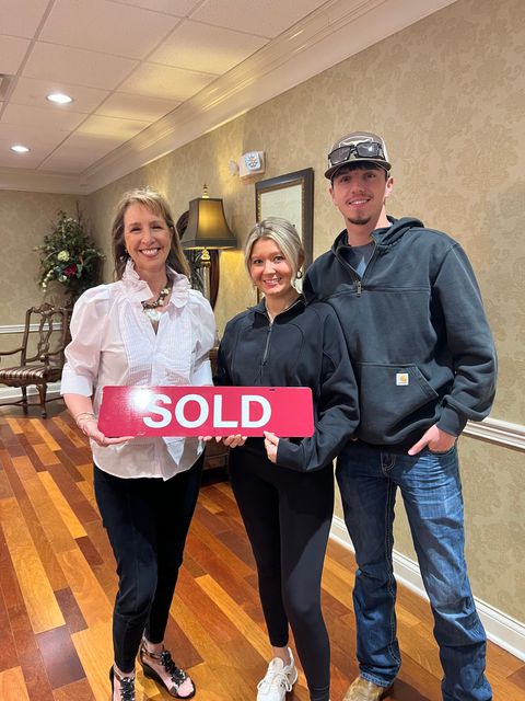 Three people indoors holding a red SOLD sign, smiling in a hallway.