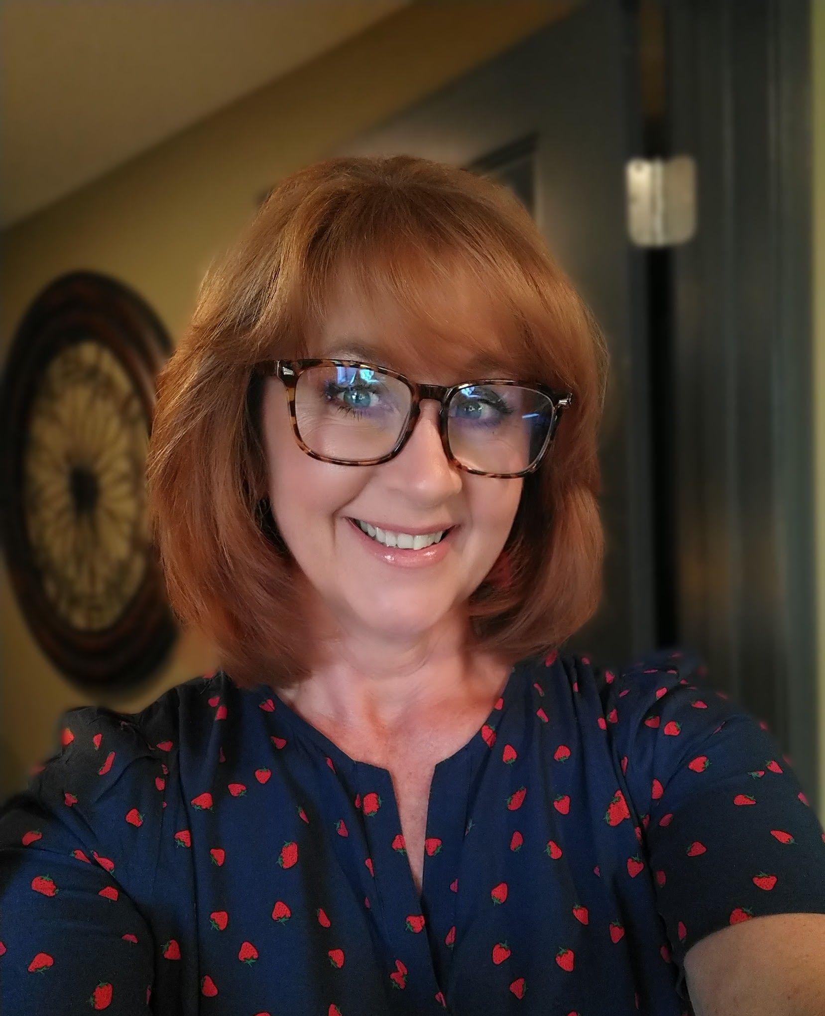 A smiling person with auburn hair and glasses wearing a blue shirt with a red floral print, standing indoors.