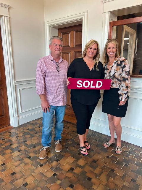 Three people holding a red “SOLD” sign in a bright house interior