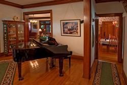 A black grand piano sits in a hardwood-floored room, with a view through doorways into a sunroom and a dining area.