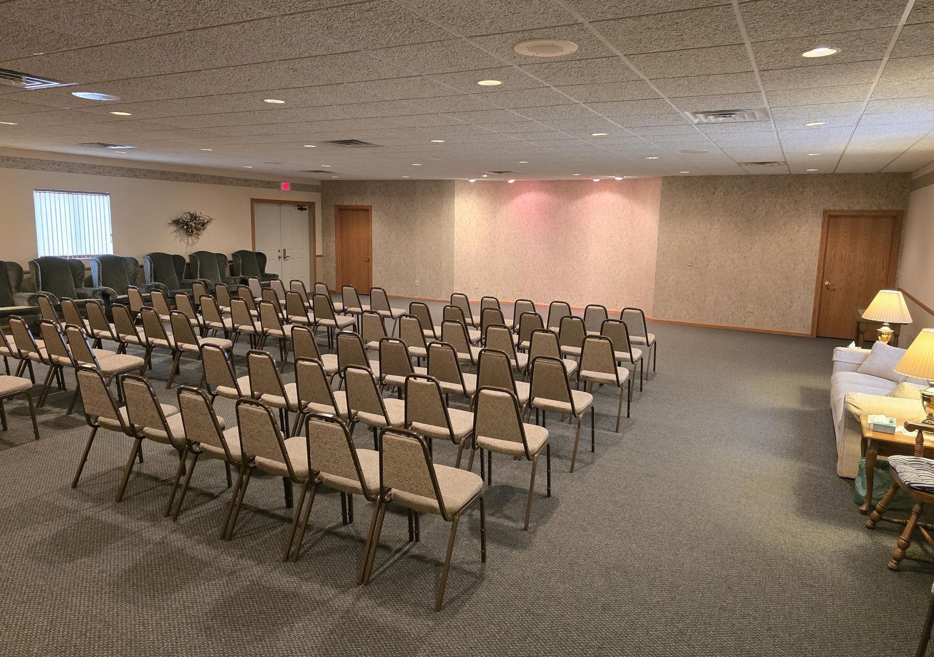 Rows of chairs facing a blank wall in a carpeted conference room with side doors and a corner seating area.