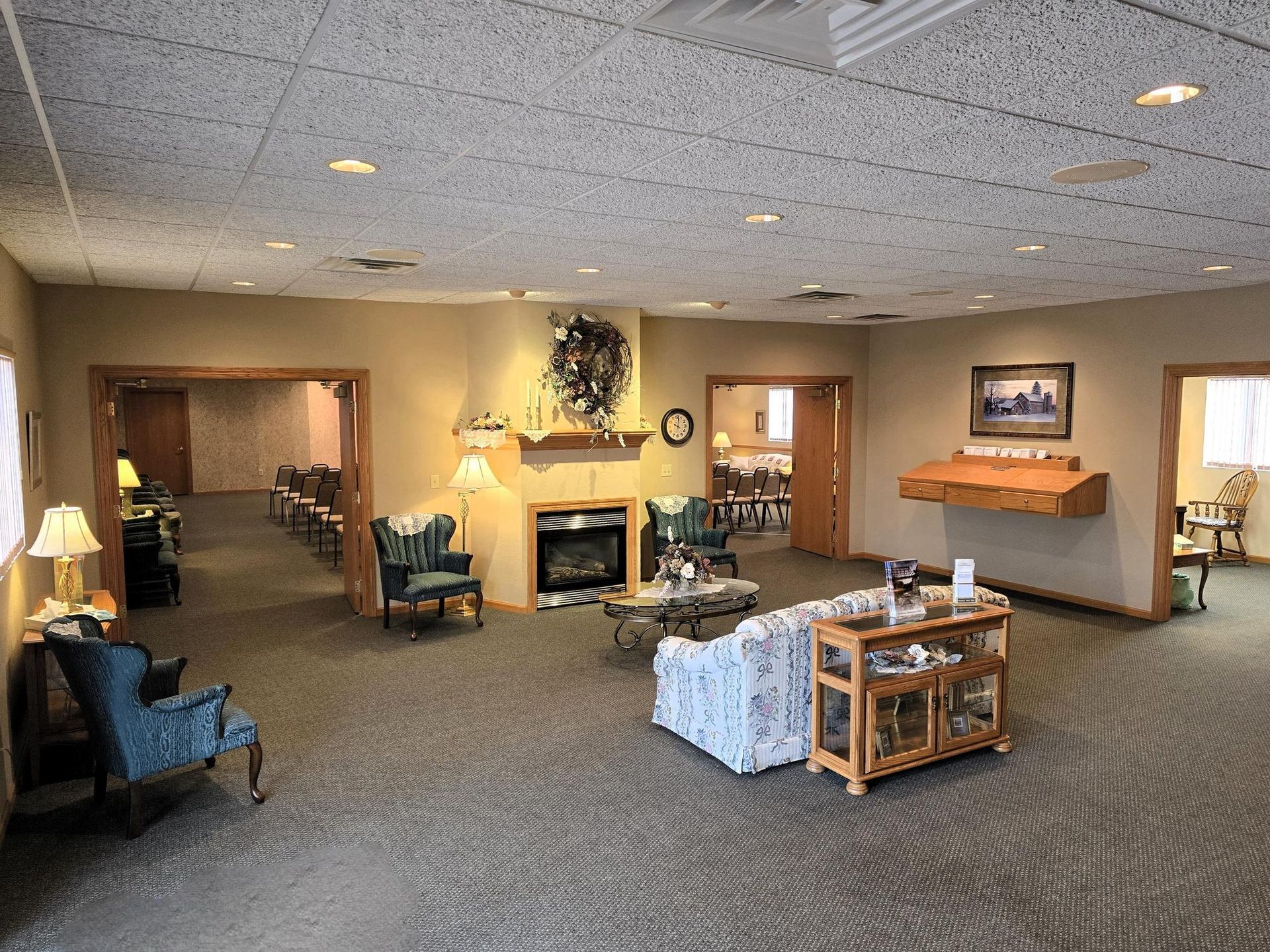A quiet, carpeted waiting area with armchairs, a central fireplace, a patterned couch, and side tables in a neutral room.