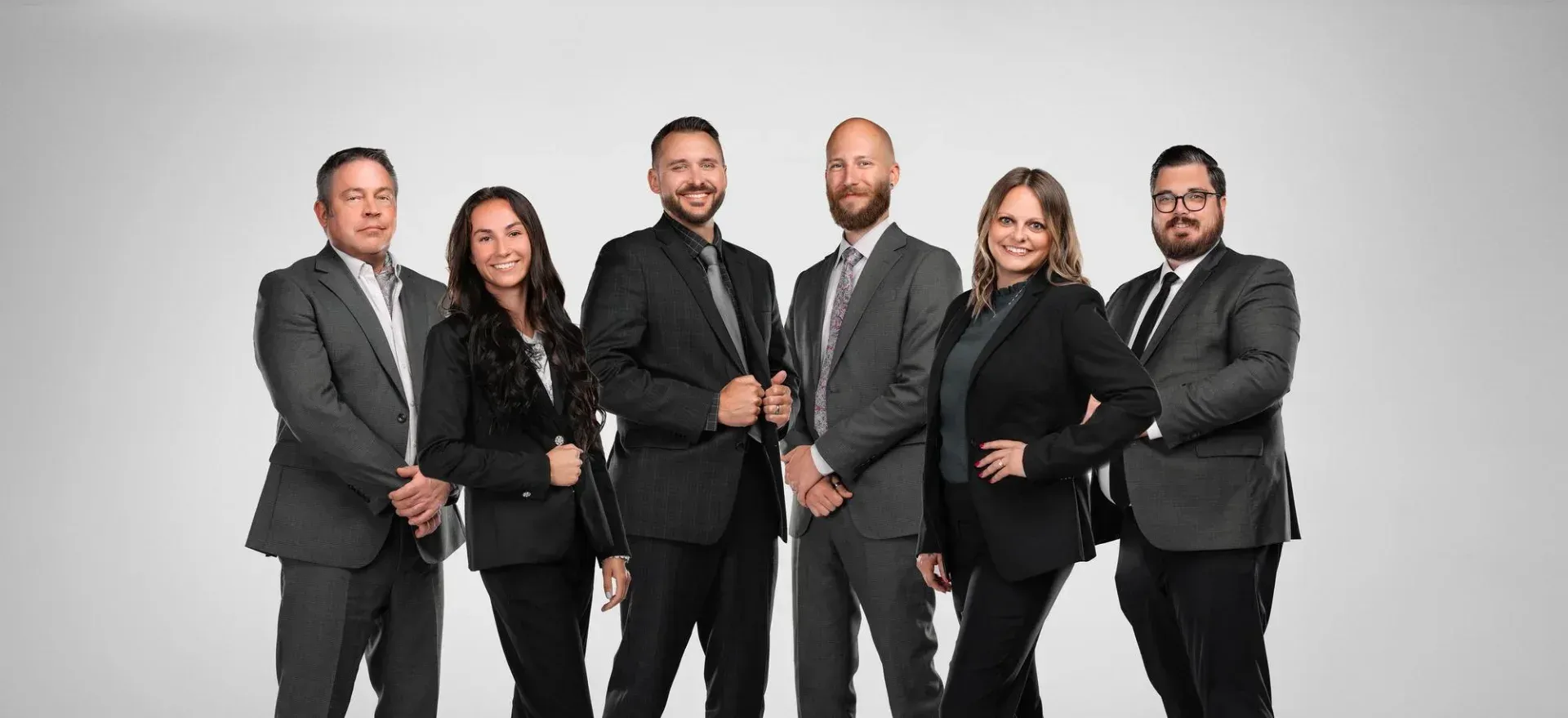 Six professionals in business attire stand in a row against a plain light gray background, smiling at the camera.