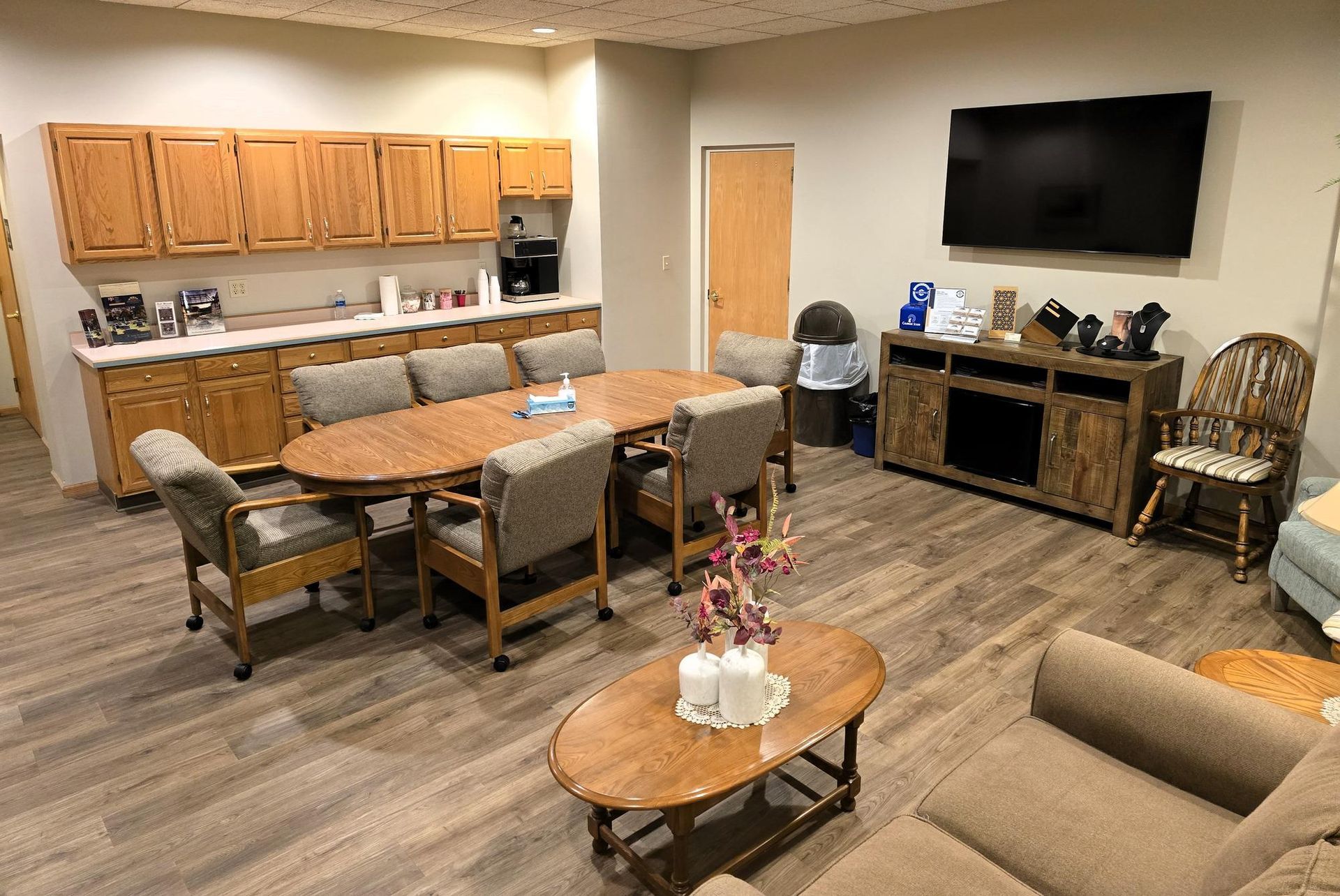 A bright breakroom with a wooden table, chairs, cabinets, a mounted television, and a seating area with a sofa.