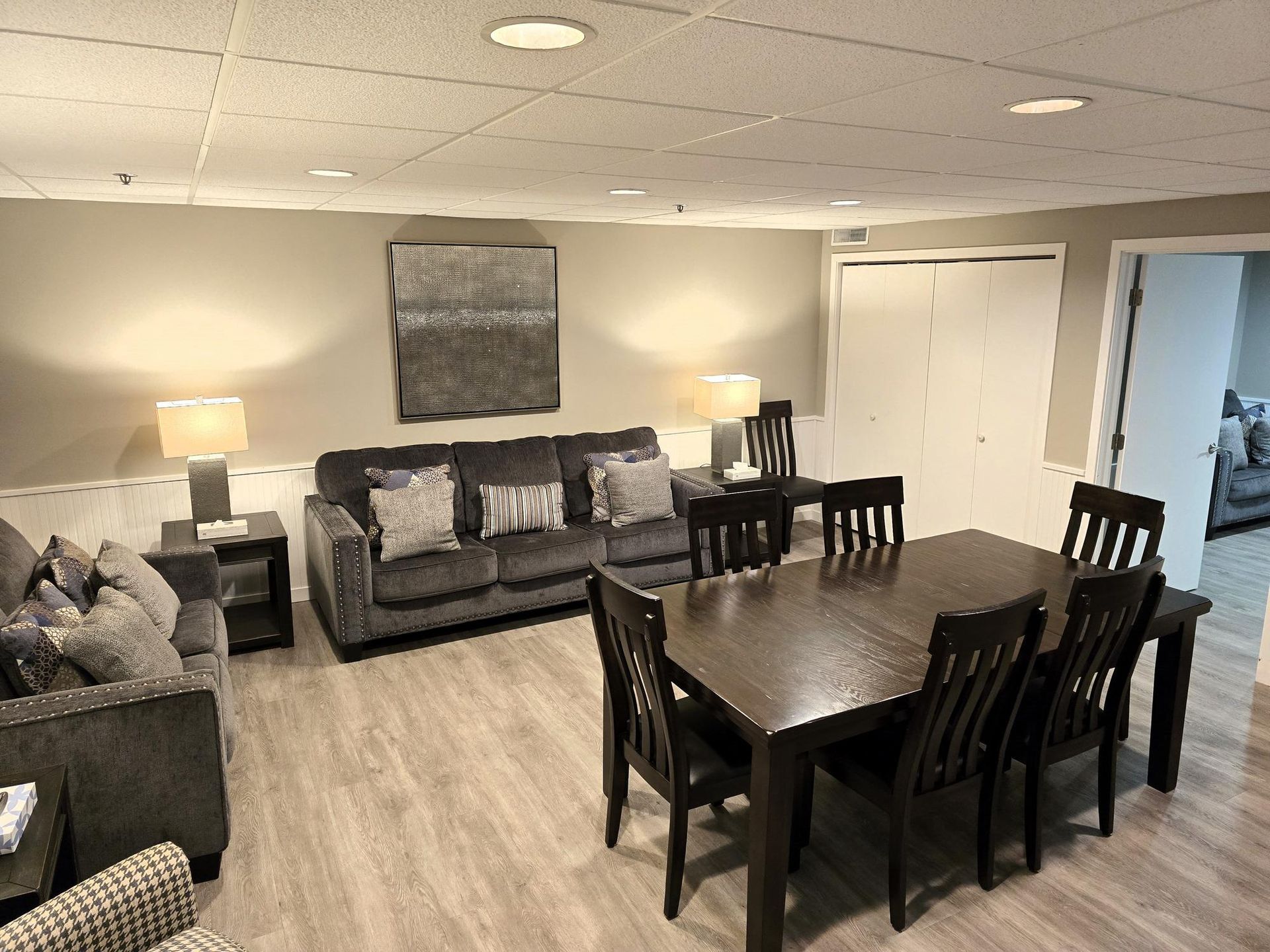 A furnished room featuring a grey couch, a dining table with six chairs, beige walls, and light wood-patterned flooring.