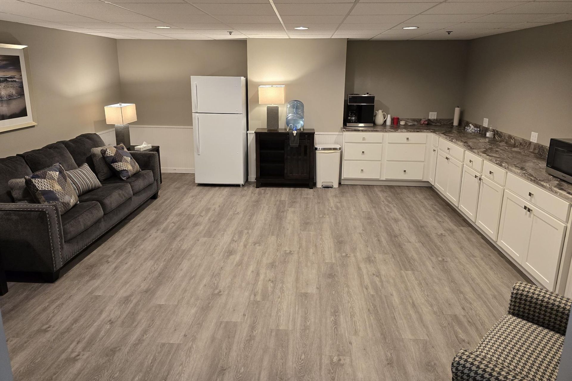 A basement living area with a dark gray sofa, white cabinets, a refrigerator, light wood floors, and neutral walls.