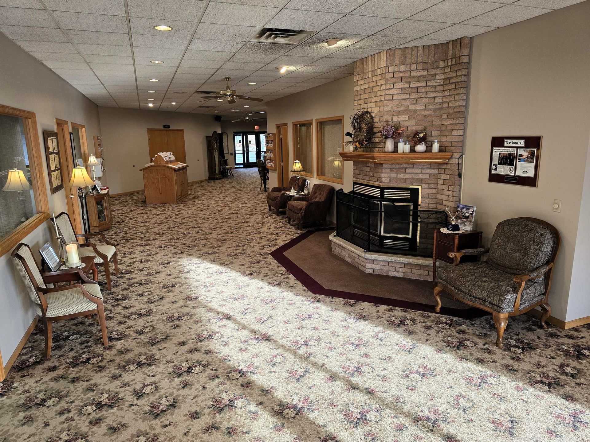 A waiting room with patterned carpet, a brick fireplace, patterned armchairs, and a distant doorway.