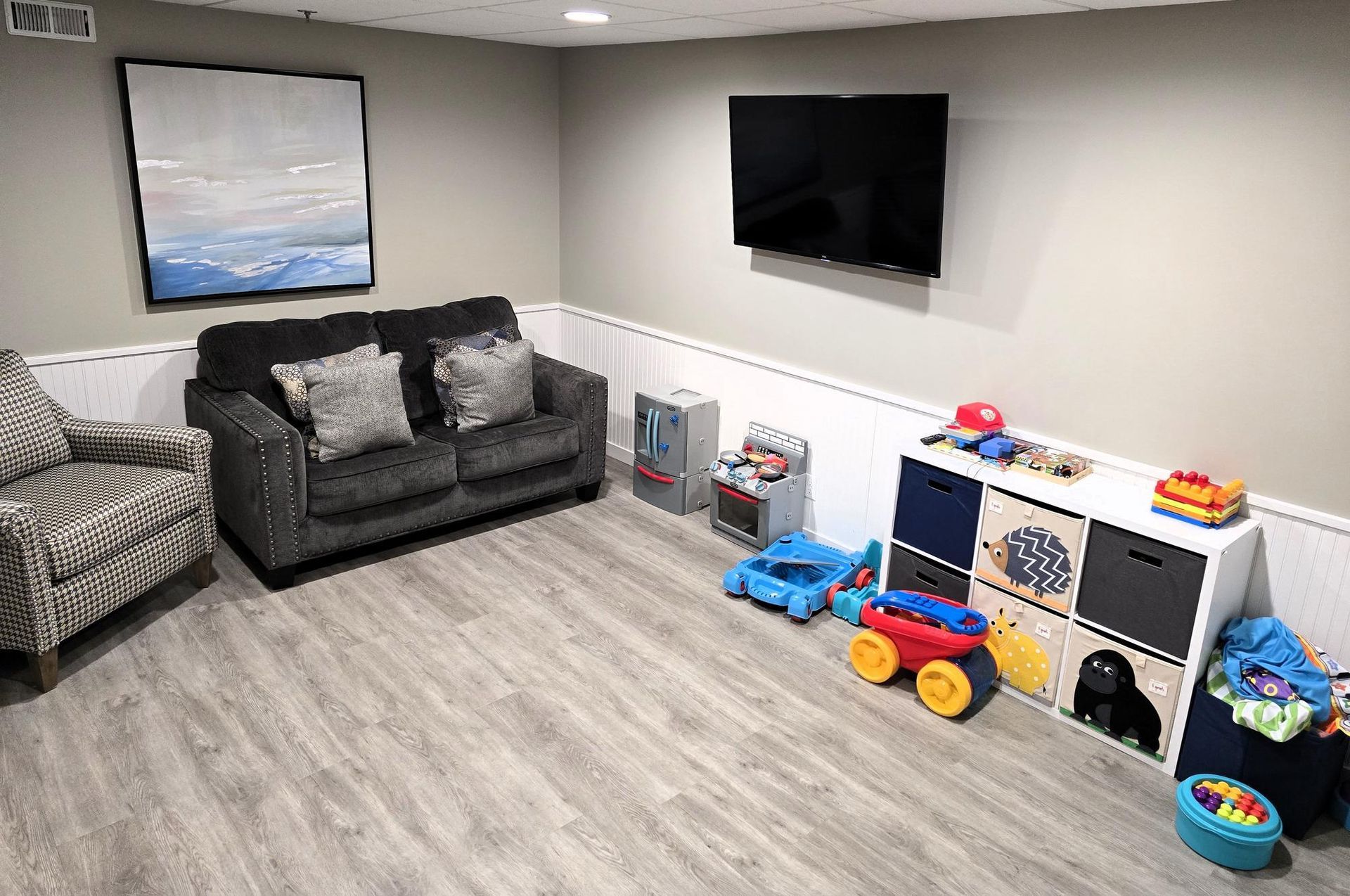 A cozy basement play area with a grey sofa, an armchair, a wall-mounted TV, and a white storage unit full of colorful toys.