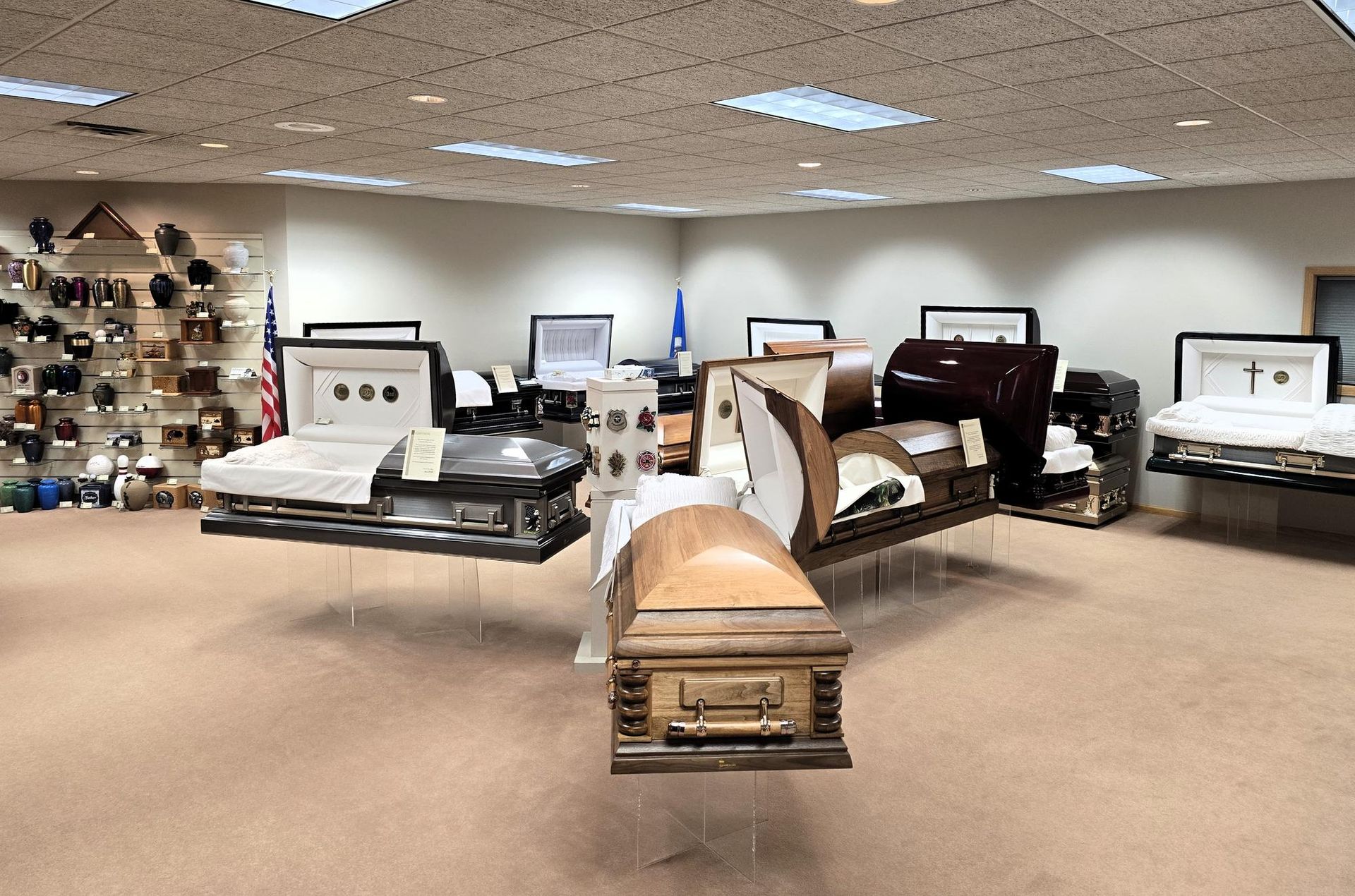 A showroom featuring various open and closed caskets arranged on pedestals, with a display of urns on a wall in the back.