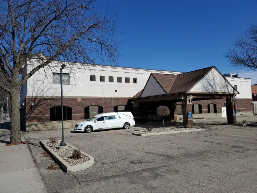 A white hearse parked in front of a two-story brick and stucco building with a prominent covered entrance.