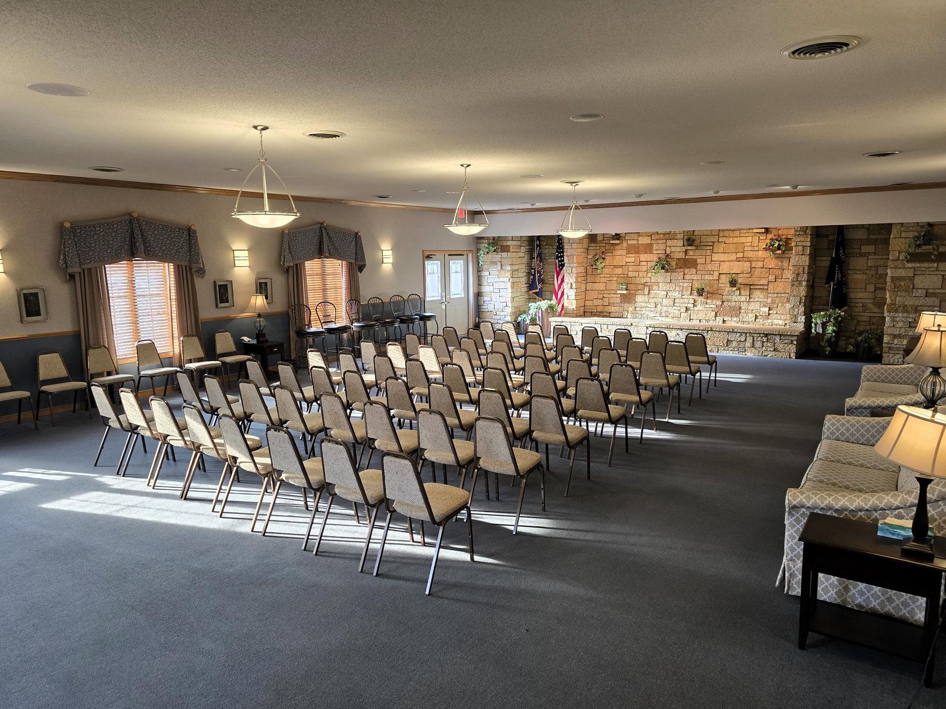 Rows of chairs face a stone wall and stage in a large, carpeted event space with pendant lighting.