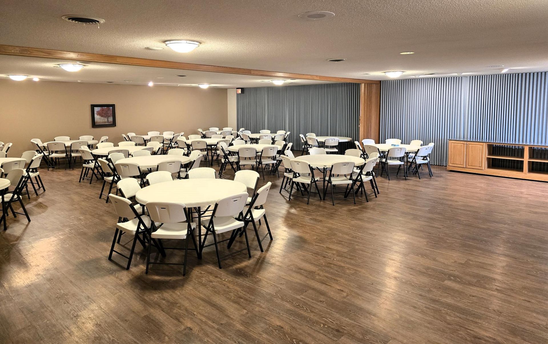 A large event space with round tables and white folding chairs arranged on a wooden floor under recessed lighting.