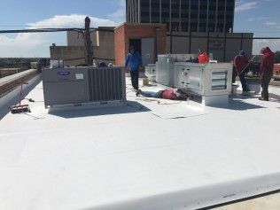 Workers service HVAC units on a white flat roof against a bright blue sky.