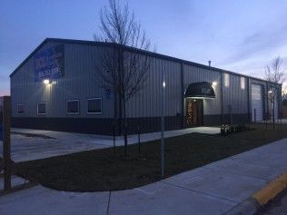 A modern gray metal commercial building at dusk with a covered entrance, large windows, and a paved sidewalk in front.