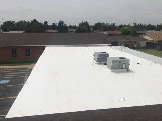 A flat, white-roofed building with two HVAC units on the surface, overlooking a parking lot and neighborhood homes.