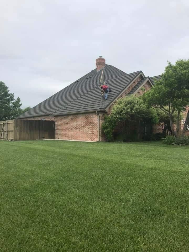 A worker in a red shirt stands on the sloped, shingled roof of a brick house next to a large tree on a lawn.