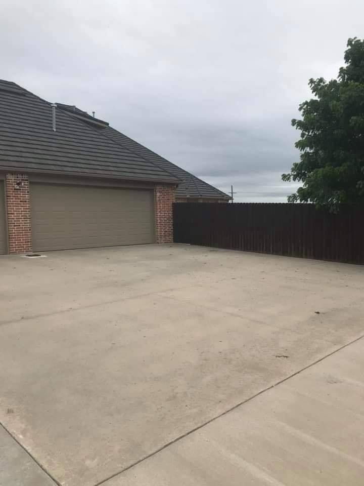 A brick house with a tan garage door sits at the end of a wide concrete driveway next to a tall wooden privacy fence.