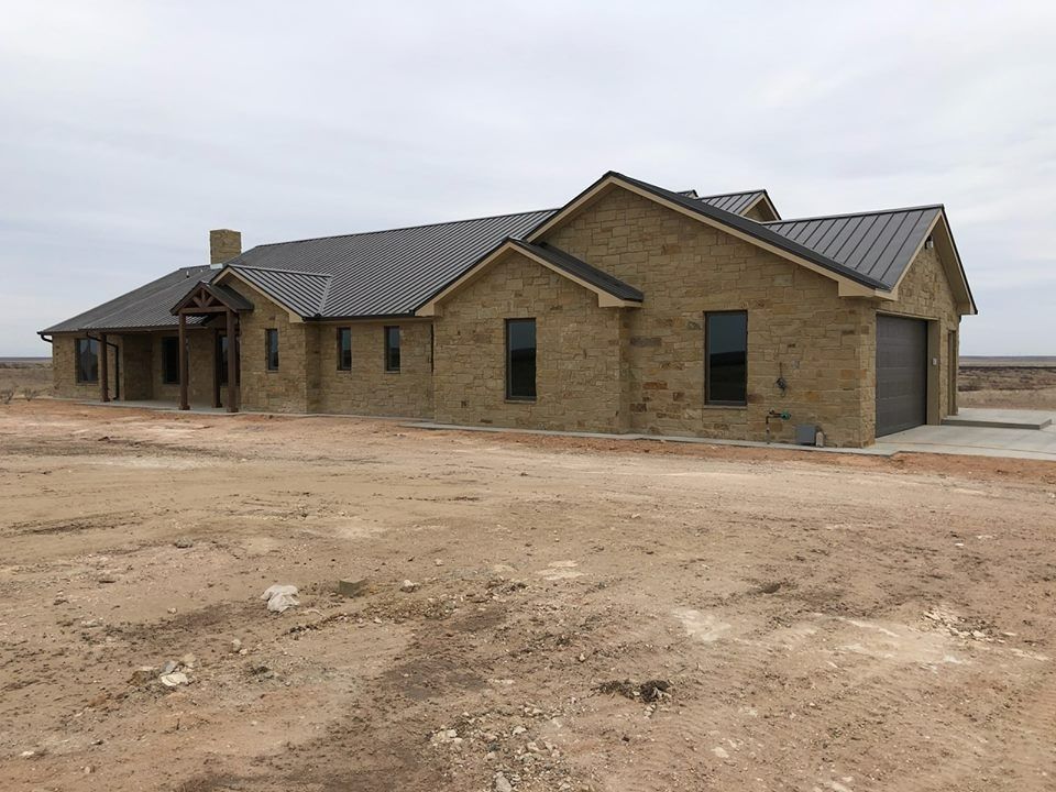 A newly constructed, single-story light brown stone house with a dark metal roof sits on an expansive, barren dirt lot.