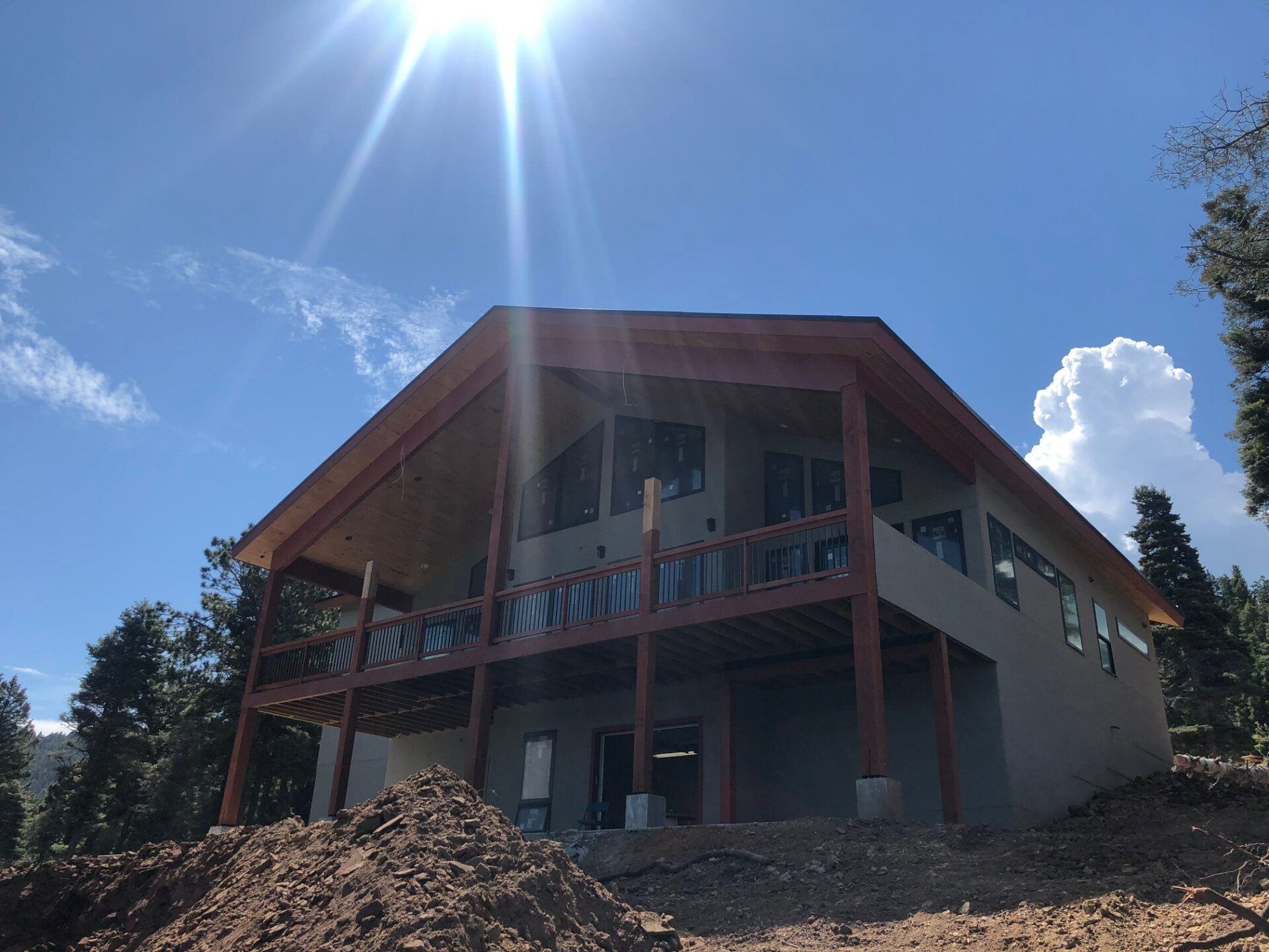 A two-story house with a wooden deck under construction, set in a sunny, rural area with dirt landscaping and trees.