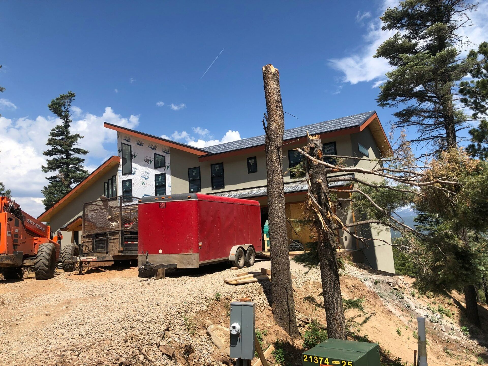 A large house under construction sits on a dirt lot with a red trailer and orange construction equipment in the foreground.
