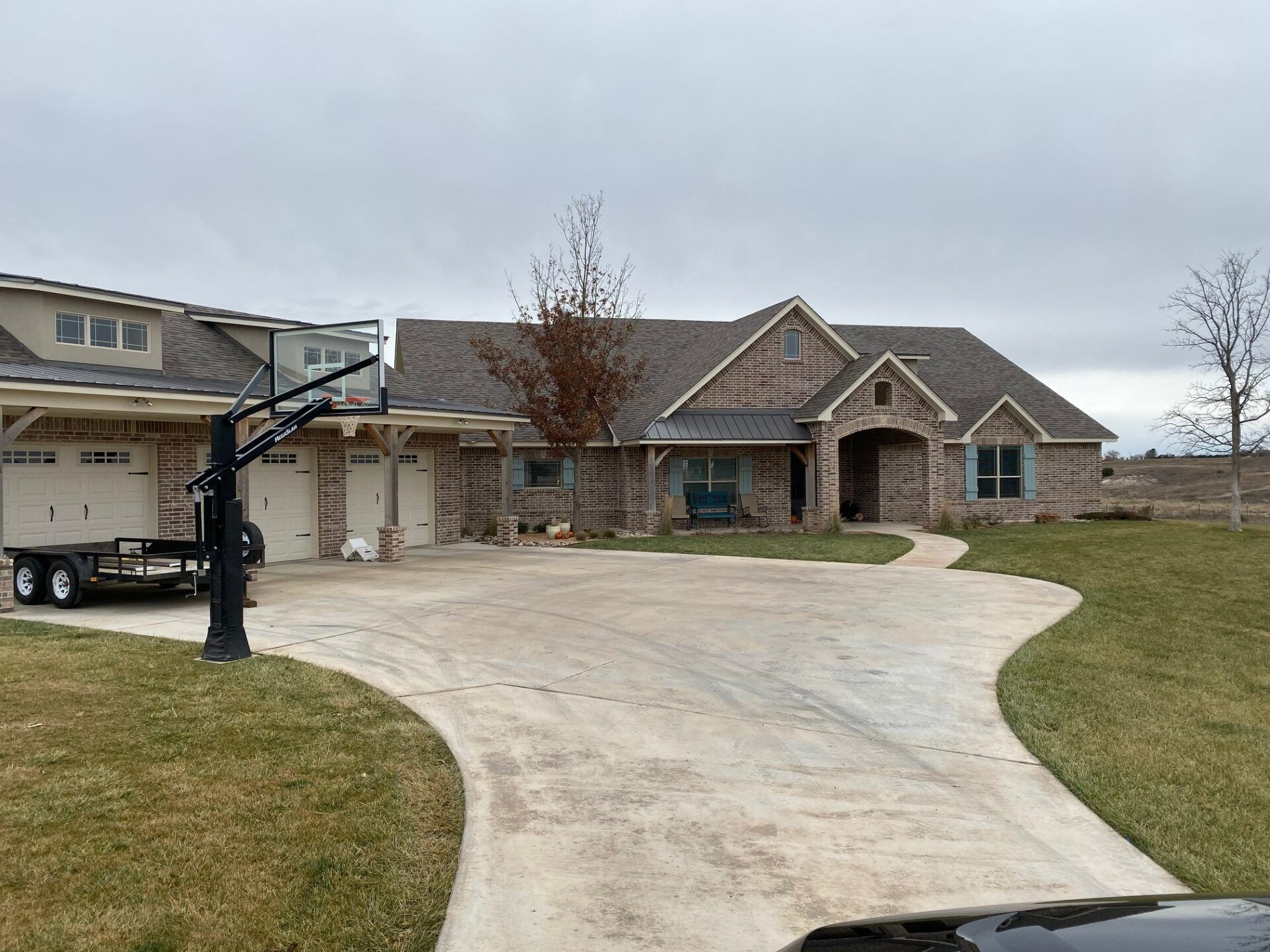 A large brick house with a multi-car garage, a wide concrete driveway, and a basketball hoop under a cloudy sky.