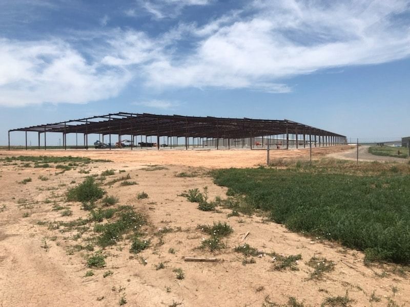 A wide, dirt lot featuring a large, dark steel framework of an unfinished warehouse building under a blue, cloudy sky.