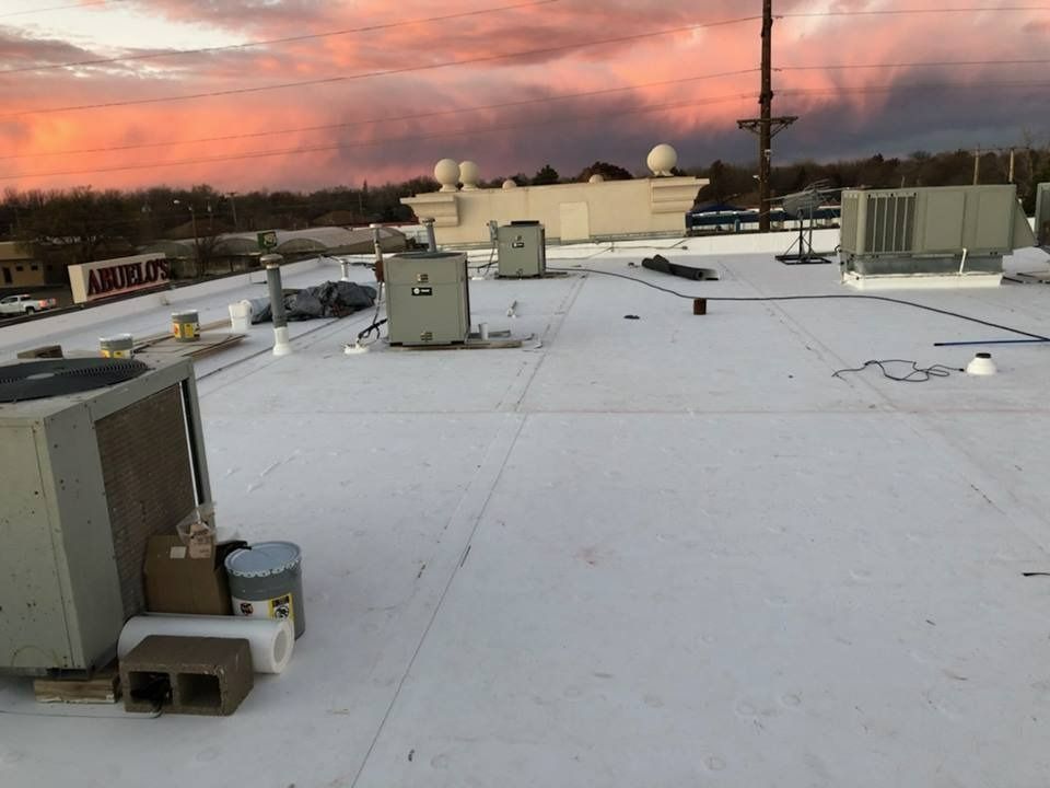 A rooftop with white membrane roofing, HVAC units, and materials, set against a vibrant pink and orange sunset sky.
