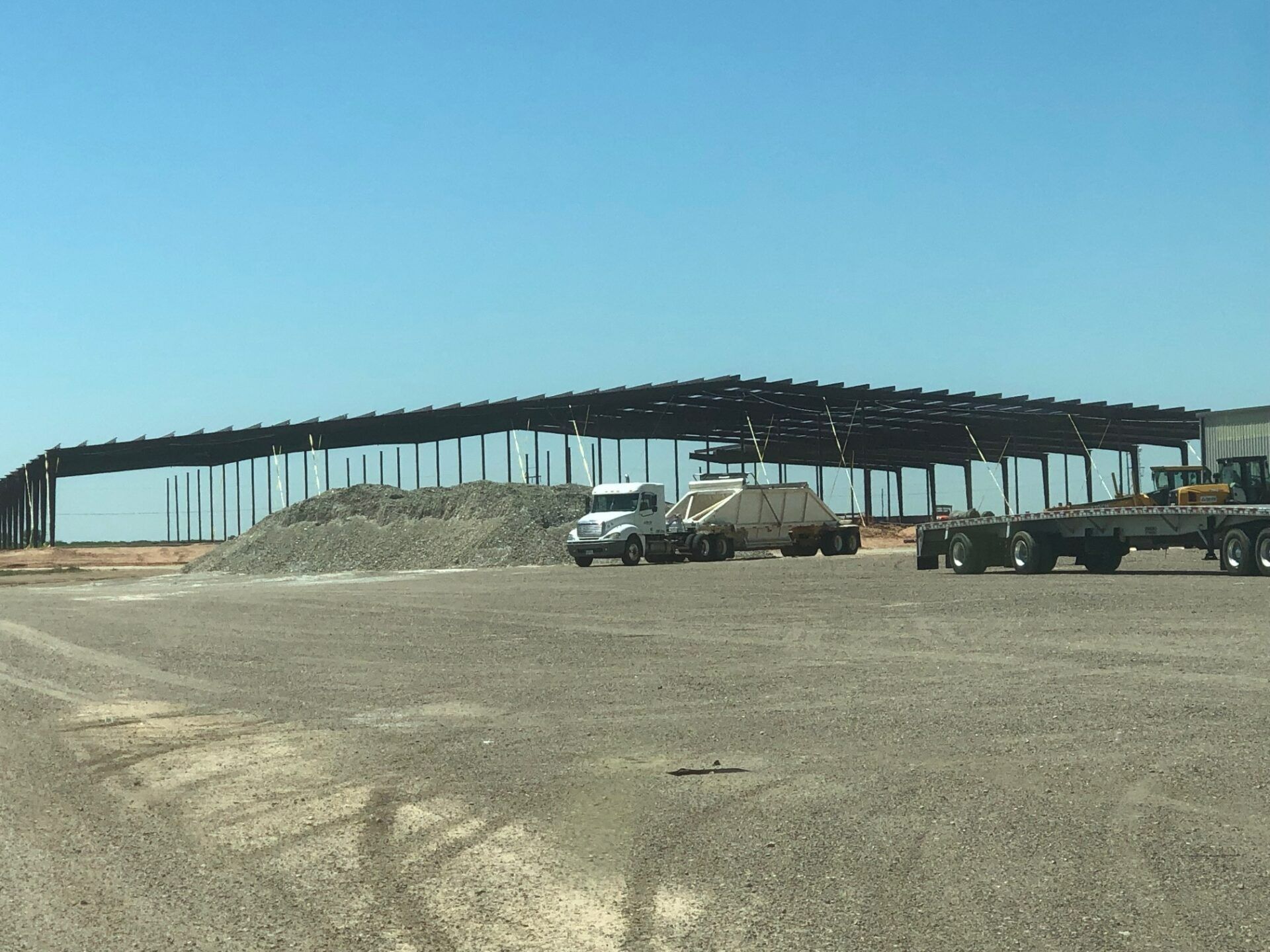 A construction site showing a large metal building frame, a gravel pile, and heavy trucks under a clear blue sky.