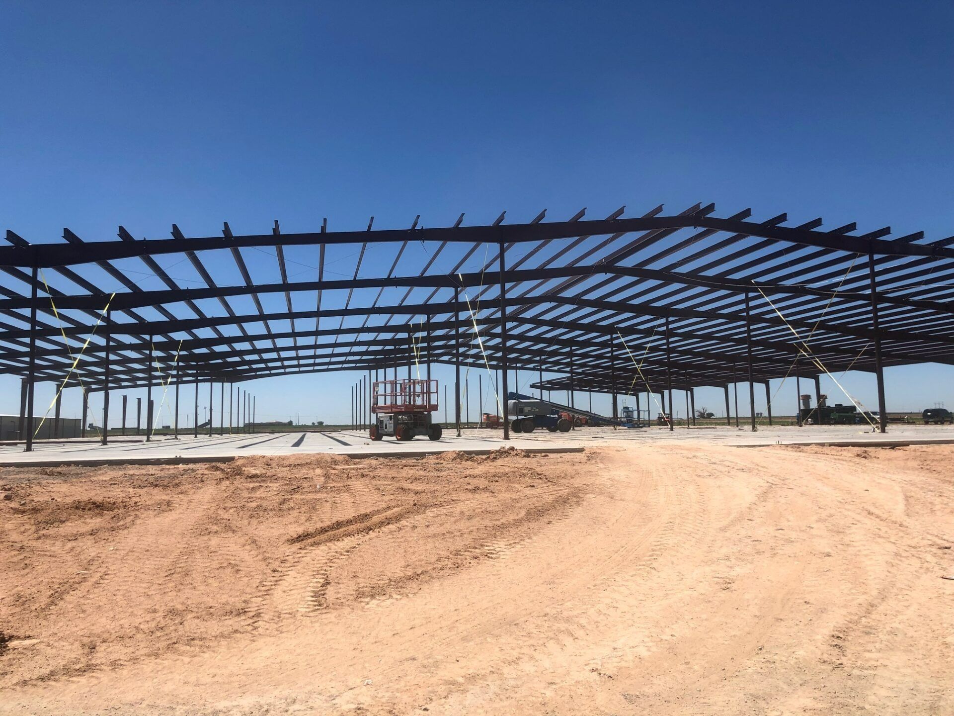 A wide view of a steel frame structure under construction on a dirt lot against a clear blue sky.