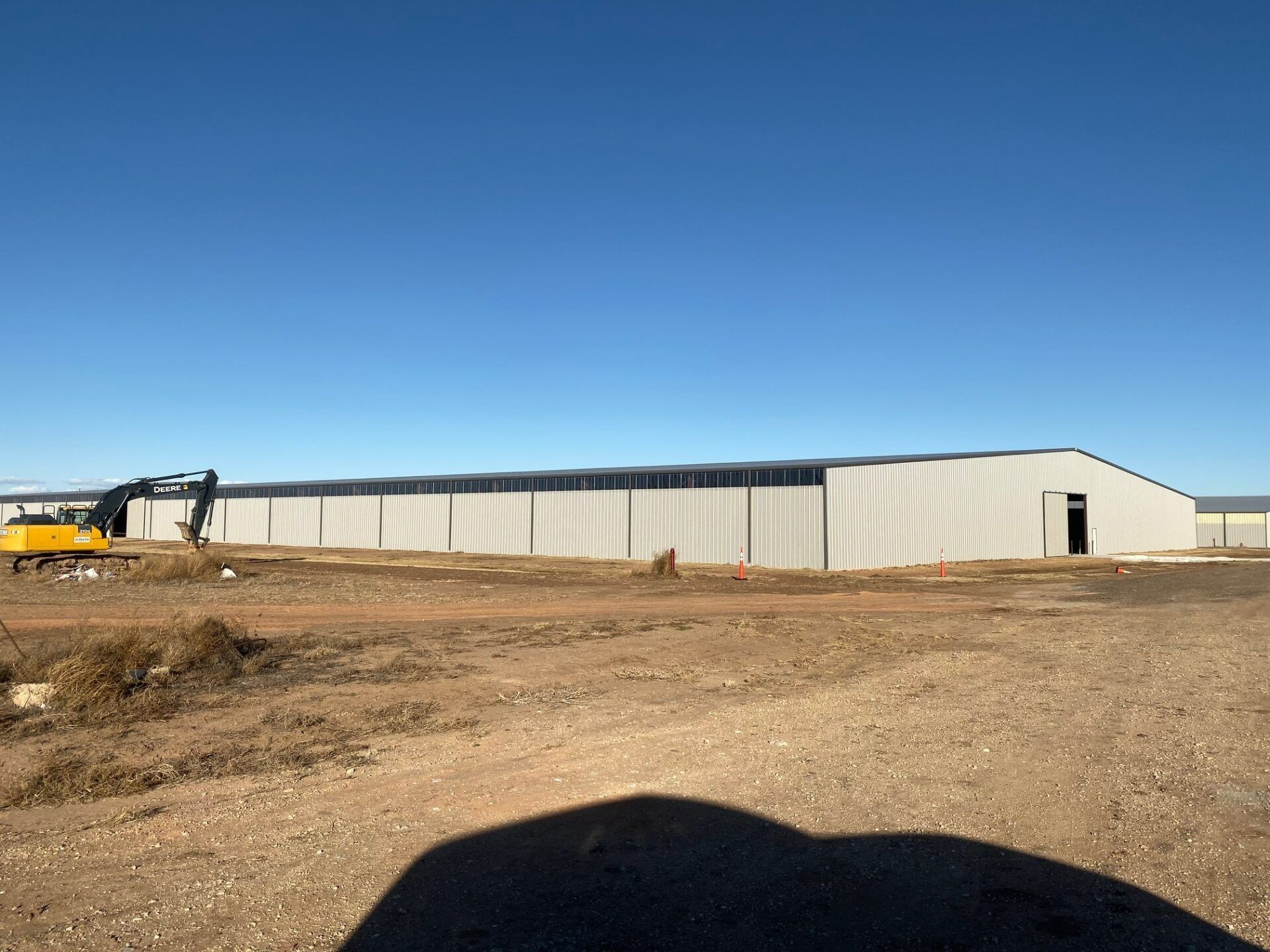 A large, long metal industrial building sits on a dirt site under a clear blue sky, with a yellow excavator to the left.