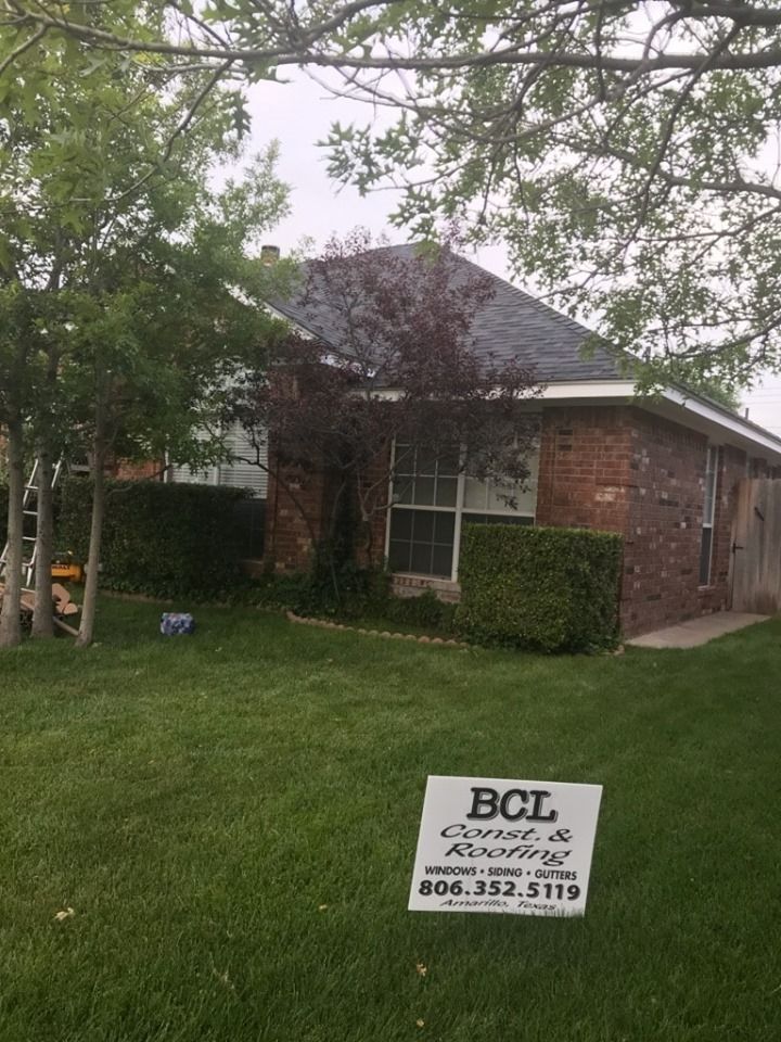 A one-story brick house with a dark shingle roof stands behind a lawn with a BCL Construction & Roofing sign in front.