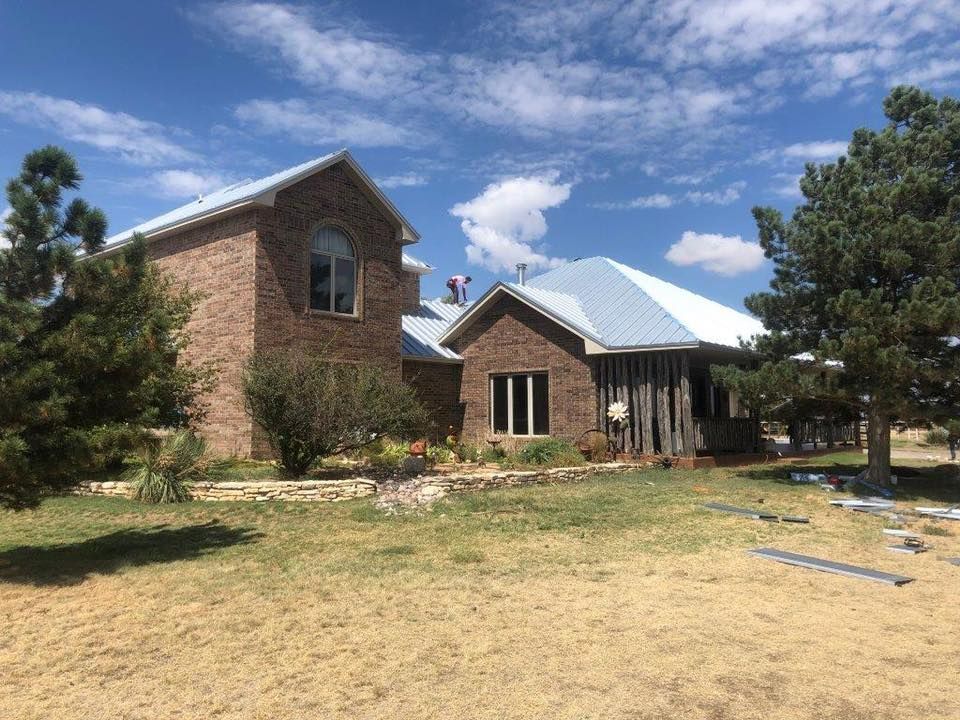 A brick house with a newly installed light-colored metal roof under a bright, sunny sky with scattered clouds.