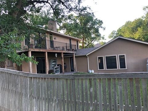 A two-story tan house with a wooden deck and attached garage, viewed over a wooden fence in a treed yard.