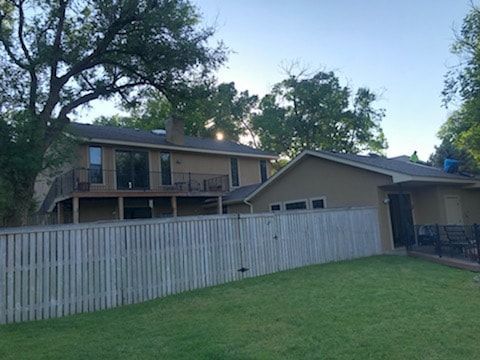A two-story tan house with a large wooden deck behind a gray wooden fence in a grassy backyard under a clear sky.