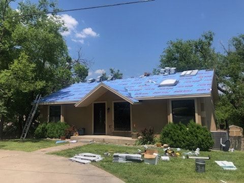 A beige, single-story house undergoing roof renovations, featuring a blue protective underlayment on the roof.