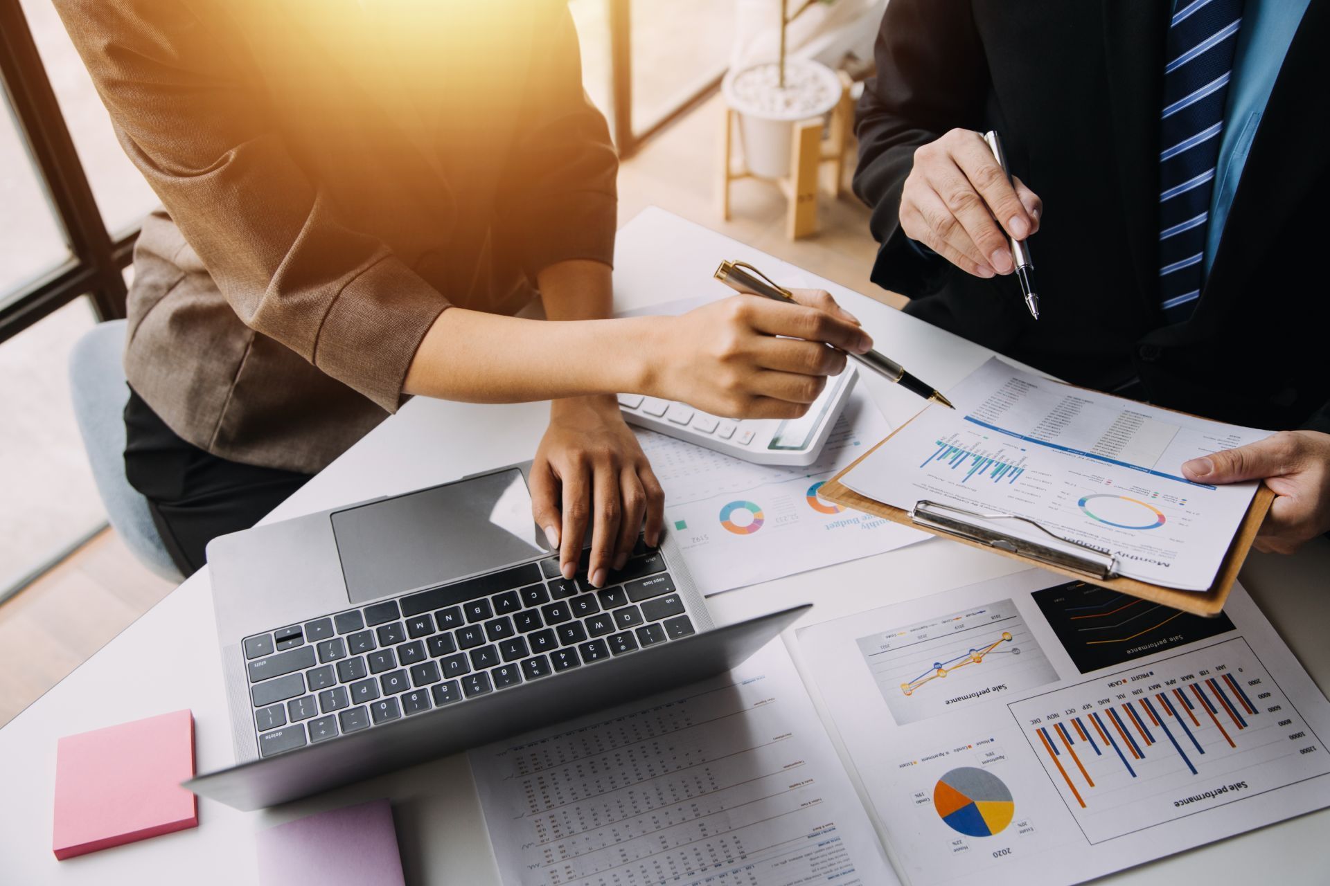 Two businesspeople reviewing documents, one using a laptop, charts, and calculator on a desk.