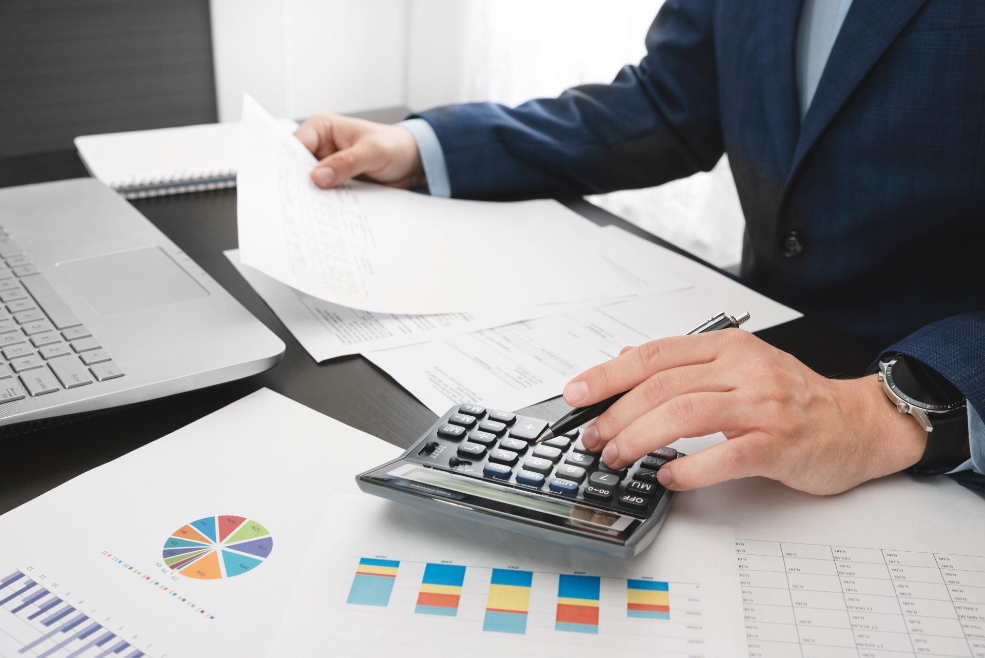 A person in a suit using a calculator while reviewing financial documents, a laptop, and charts on a desk.