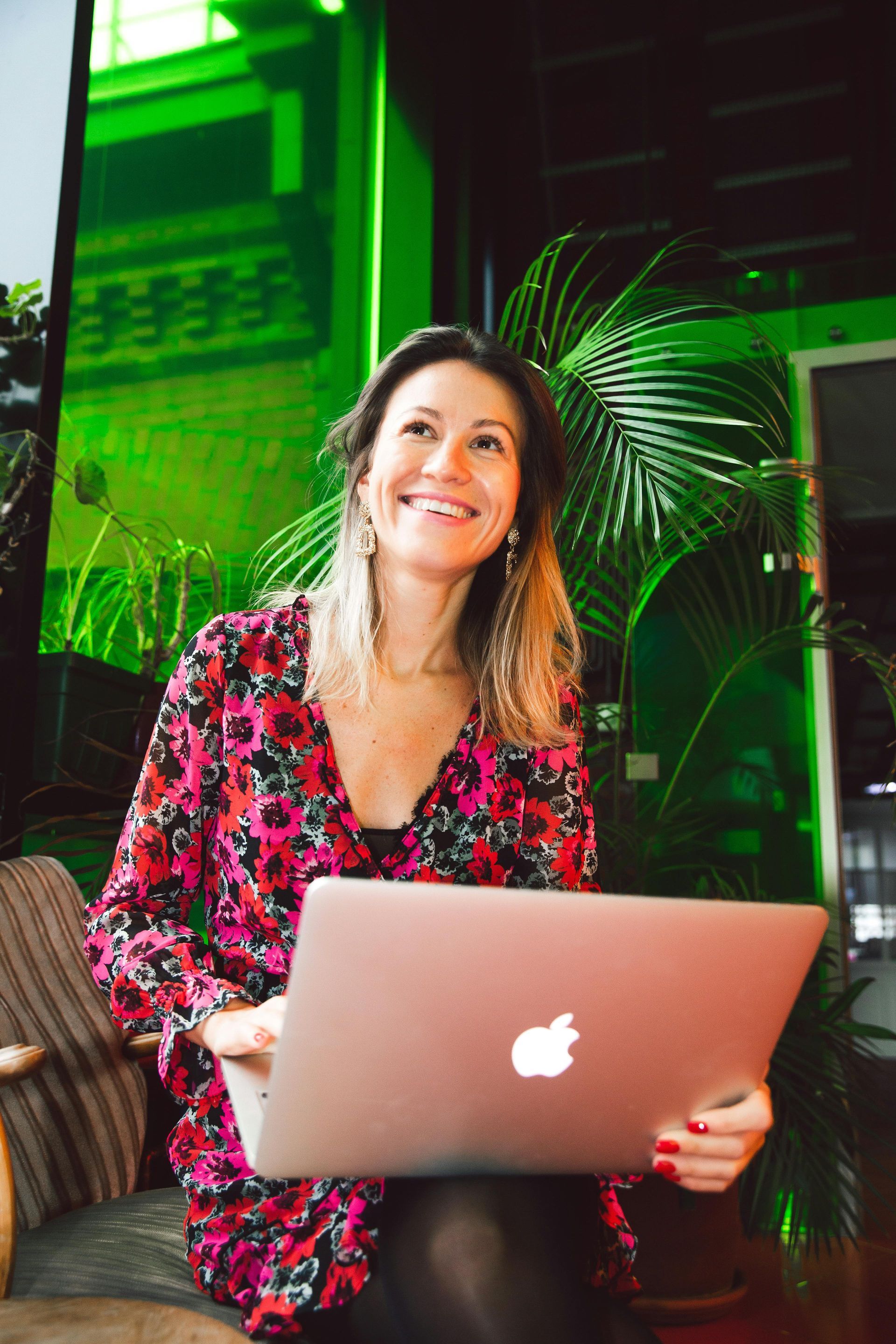 Woman using laptop, smiling. Wearing floral dress, sitting near green plants.