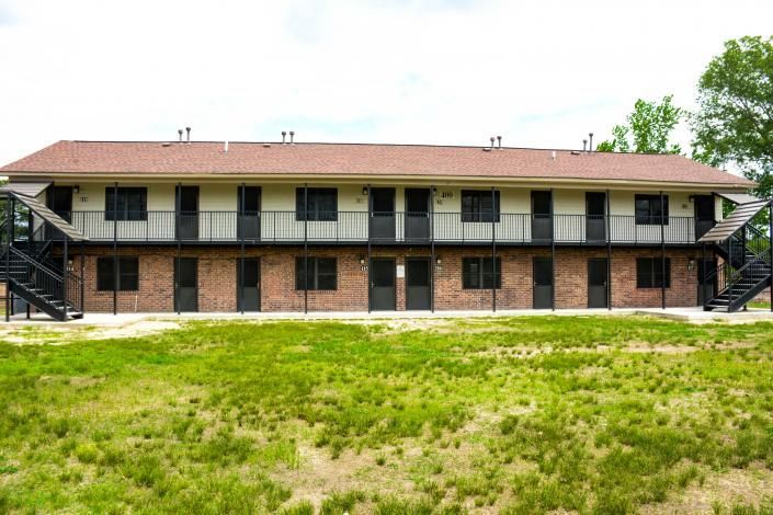 Two-story brick motel with exterior staircases and a red roof, set behind a grassy field