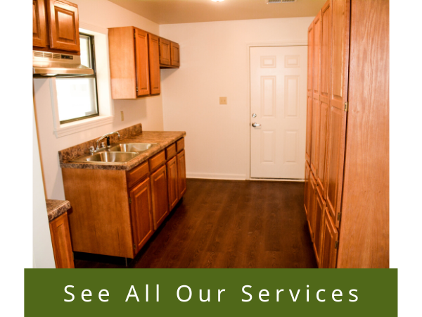 Small kitchen with wood cabinets, sink, white door, dark floor, and “See All Our Services” banner at bottom