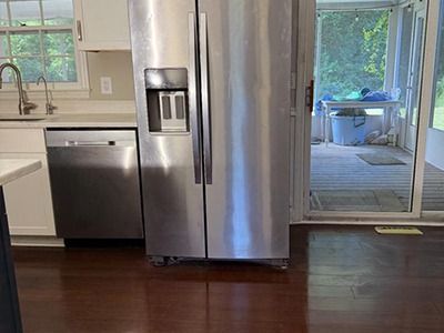 A stainless steel refrigerator stands next to a dishwasher in a kitchen, with a view of a porch through a sliding glass door.