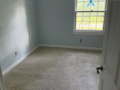 An empty bedroom with light blue walls, beige carpeting, and a white-trimmed window with a blue cross on the glass.