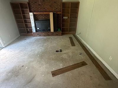 A room with a brick fireplace, built-in wooden shelves, and a few dark brown vinyl flooring planks on a concrete subfloor.